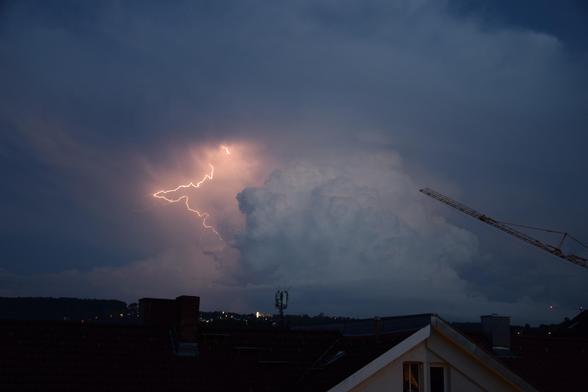 Gewitterwolke am Horizont mit einem rot-leuchtenden Blitz links daneben. Am unteren Bildrand sind die Häuserdächer Stuttgarts zu sehen.