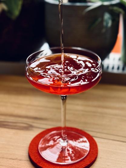 a bright red cocktail being poured into a chilled coupe with a dynamic reflective swirling surface to the liquid in the glass