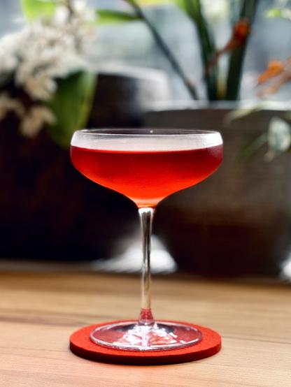 a bright red cocktail in a frosty coupe sitting on a wooden table with potted plants in the background