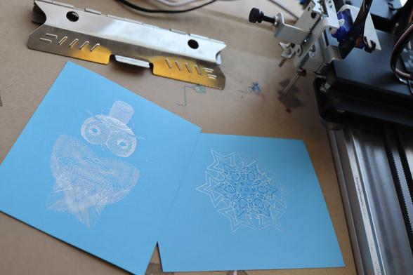 Two plots on blue cardstock, on the plotting surface by my AxiDraw: a CAD collage of my robot owl Archimedes, and a blue-white-silver snowflake.