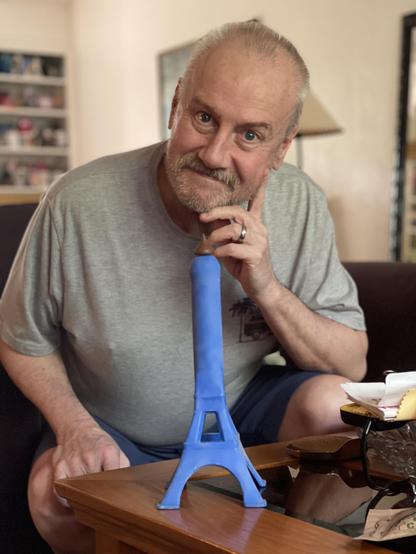 A man sits behind a coffee table. On the coffee table is a blue dildo in the form of the Eiffel Tower. On top of the dildo is a Hershey’s Kiss.