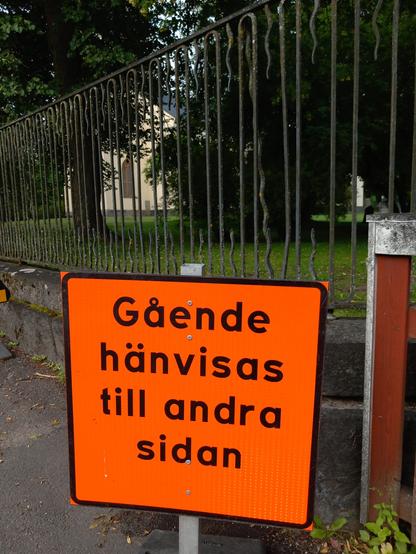 Orange vägskylt, "Gående hänvisas till andra sidan", vid staketet till Avesta kyrkas kyrkogård