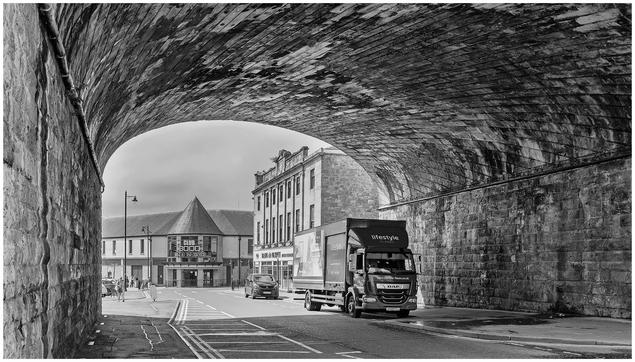 A black and white image depicting a street scene under an archway. A truck is parked on the right, while another vehicle drives past. In the background, a building with a triangular roof features signage for a bingo club.