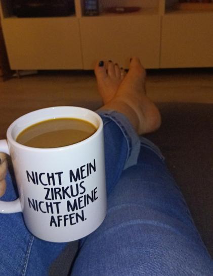 Zwei Beine in Jeans, barfuß, de Beine hochgelegt auf einem Sofa.
Die linke Hand hält einen weißen Kaffeebecher mit der Aufschrift 'Nicht mein Zirkus, nicht meine Affen'