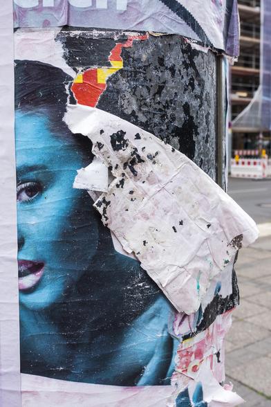 colour photograph, portrait format. a traffic light column, on it a half blue face, left and bottom white ‘framed’, half torn off poster, construction site in the background.

farbfotografie, hochformat. eine ampelsäule, darauf ein halbes, blaues gesicht, links und unten weiß "gerahmt", halb abgerissenes poster, baustelle im hintergrund.