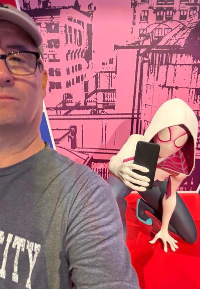 David Rider’s Spidey-sense is tingling because the costumed heroine Spider-Woman AKA Ghost-Spider AKA Spider-Gwen, who is crouched behind him, is taking a photo of the indie author who wrote her favorite novel Anca’s Undead Playlist (a vampire story about a cursed teenage musician, much like herself)