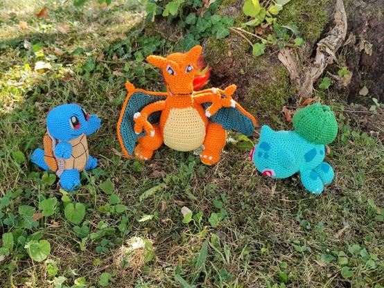 Three crocheted Pokémon characters—Squirtle, Charizard, and Bulbasaur—sit on a grassy patch outdoors. Squirtle, on the left, is blue with a brown shell; Charizard, in the center, is orange with wings and a flame-tipped tail; and Bulbasaur, on the right, is green with a bulb on its back. The handmade figures are crafted in amigurumi style, and surrounded by natural textures like grass, leaves, and a tree trunk, adding charm to their woodland setting.