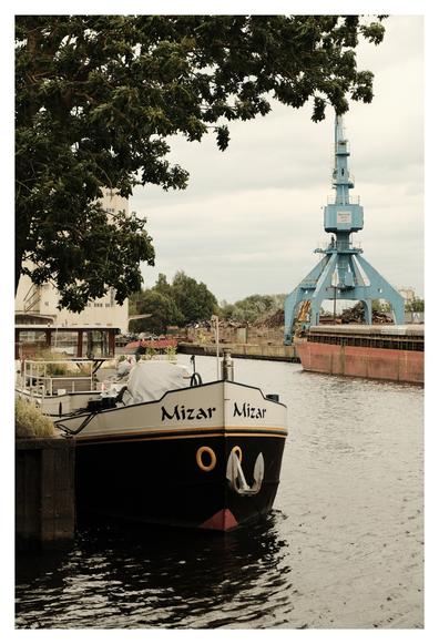Ein Schiff mit dem Namen „Mizar“ liegt am Kai eines Hafens. Im Hintergrund steht ein großer blauer Kran, daneben ein rostiges Frachtschiff. Links ragt ein Baum ins Bild, am Ufer wachsen Pflanzen.
