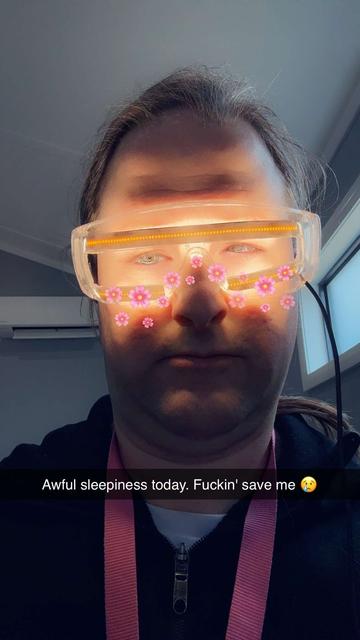 A girl with pretty eyes looking beyond tired. Pink Snapchat filter flowers adorn her nose and cheekbones. She is wearing a thick pink lanyard and a black zippered jacket. Indoors. Dreary day. Her eyes are wrapped in modified safety glasses, with high-power COB LED strips facing inward, top and bottom. Caption reads: awful sleepiness today. Fuckin' save me. 😢