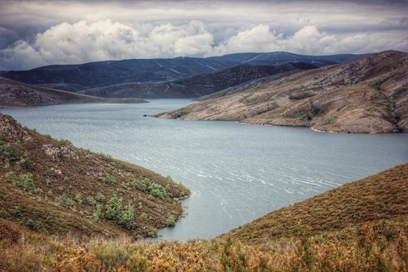 A wide shot of a tranquil lake nestled between rolling, reddish-brown hills under a dramatic cloudy sky. The water is calm with subtle ripples, reflecting the light. Green foliage is visible on the hillsides, particularly on the left, adding a touch of vibrancy to the earthy tones of the landscape. Distant mountains fade into the hazy background.