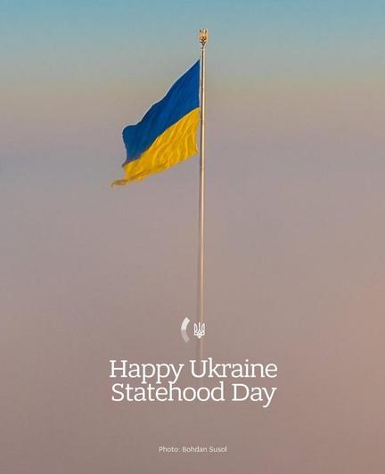 The image depicts the flag of Ukraine flying on a flagpole against a backdrop of a gradient sky transitioning from blue to a soft pinkish hue. The flag consists of two horizontal stripes: the top stripe is blue, and the bottom stripe is yellow. At the bottom of the image, there is text that reads "Happy Ukraine Statehood Day." Additionally, there is a small emblem or logo featuring the Ukrainian Tryzub (trident) above the text. The photo credit is given to Bohdan Susol.