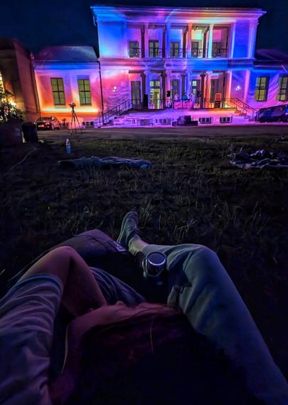 A low-angle shot from the perspective of someone lying on the grass, looking towards a grand, illuminated white mansion at night. The mansion is bathed in shifting purple, pink, and blue lights, highlighting its columns and multiple windows. Below the mansion, on a patio, figures are visible, suggesting a gathering or performance. In the foreground, the legs and torso of the person taking the photo are visible, wearing comfortable clothing and holding a vacuum flask with their legs. A child has their head in the person's lap, and only the head and left arm of the child are visible. Empty blankets are scattered on the dark grass around them, some with bottles nearby.