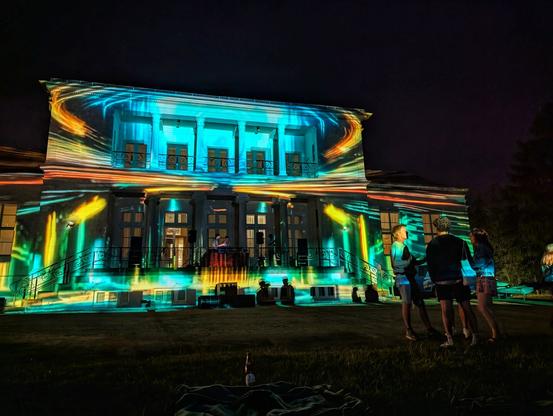A stately white mansion is brightly illuminated at night by an elaborate light projection show. Swirling patterns of orange, yellow, and turquoise light stream across its façade, highlighting the columns, windows, and balconies. In front of the mansion, several people stand on the dark grass, watching the display, with three people on a small stage near the entrance. A blanket and a bottle lie in the foreground.