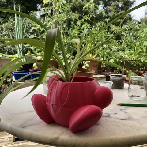 Cache pot en forme de Kirby et rose en impression 3D avec un chlorophytum dedans dont les feuilles dépassent, posé sur une table en marbre avec jardin en fond