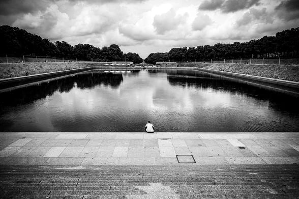 The image captures a solitary individual sitting at the edge of a large reflecting water basin. The basin dominates the foreground, its dark waters mirroring the dramatic, cloud-filled sky above. In the background, a line of trees frames the pool, with faint figures of other individuals visible along the distant edges. The person in the foreground is positioned on a paved area, with steps leading down towards the water.