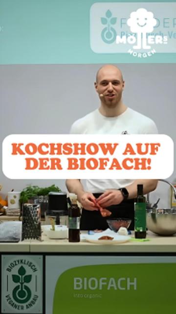 Das Bild zeigt eine Person, die eine Kochshow auf der BioFach präsentiert, einer internationalen Messe für Bio-Lebensmittel. Die Person steht hinter einem Tisch, auf dem verschiedene Zutaten und Küchenutensilien wie Flaschen mit Öl, Kräuter, Schüsseln und Schneidebretter zu sehen sind. Sie scheint aktiv am Kochen oder Vorbereiten eines Gerichts beteiligt zu sein und hält ein Mikrofon in der Hand, was darauf hindeutet, dass sie gleichzeitig spricht oder Anweisungen gibt.
Über dem Bild ist ein Text mit der Aufschrift "Kochshow auf der Biofach!" zu sehen, was den Kontext der Kochshow auf der BioFach-Messe verdeutlicht. Im Hintergrund ist ein Banner mit dem Logo "Möllers Morgen" sichtbar, was darauf hinweist, dass die Person oder die Kochshow mit diesem Betrieb verbunden ist.