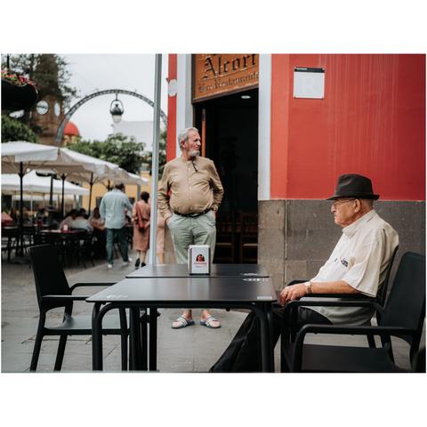 An old person is sitting on a cafeteria table outside, while another man is standing in front of the restaurant's door