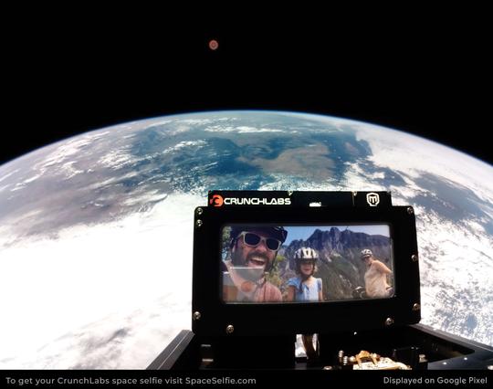 A  photo of three people wearing bike helmets in front of mount si, displayed on a screen mounted to a crunchlabs satellite with earth in the background.