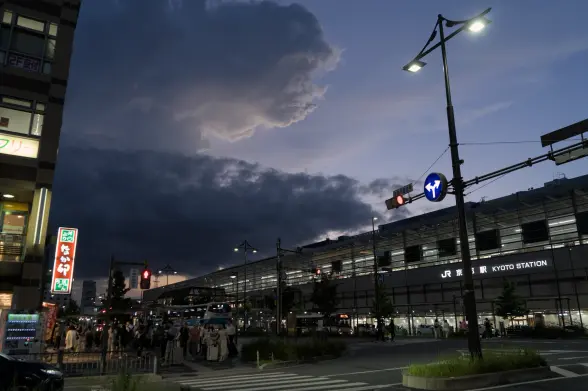 Early nightfall, low-angle photograph of a street with the large JR Kyoto train station building on the other side of the street. People are walking on the closer side of the street and several buses circulate in the street. Over the station building, a few dark-ish clouds cut into the blue sky. On the right side of the photograph in the middle of the street, a multi-purpose street lamp/trafic signpost stands.