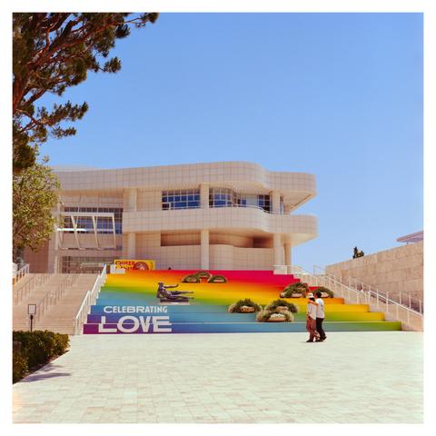 A rainbow painted steps in front the Getty Center, with the words “Celebrating Love” painted on it. A statue and some planted are placed on the steps. A couple walks by the steps.