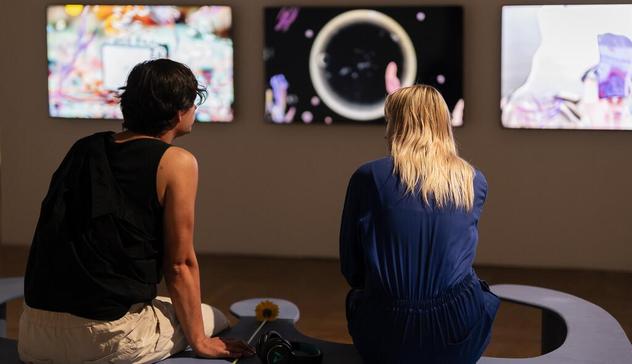 Two visitors– a man in a black short and a woman in a blue dress– sit on a bench at the IF Lab exhibition 2024/25 and watch visual multimedia projects on screens before them.