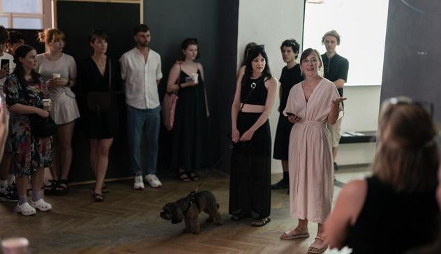 A woman in an off-white jumpsuit gives a welcoming speech to a gathering of visitors at the IF Lab exhibit 2024/25. The audience appears captivated by her speech. She stands next to a woman in a black top and trousers who has a service dog on a leash in front of her.