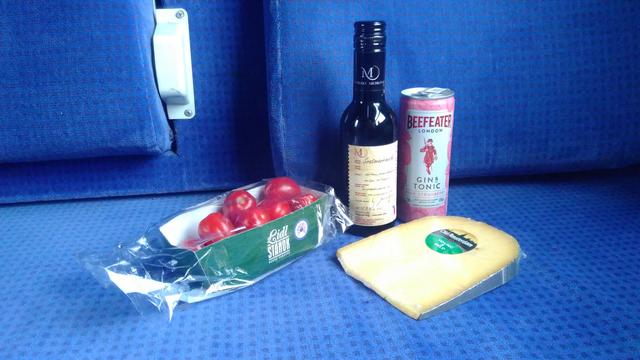 Supplies on a train carriage seat, namely a wee bottle of red wine, cheese, some tomatoes and a can of gin and tonic.