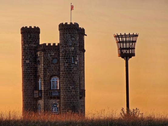 Broadway Tower, a folly in the Cotswolds, stands prominently against a soft, golden sunset sky. The stone tower with its battlements and arched windows is visible from a low angle, with tall, dry grasses in the foreground. To the right of the tower, a tall, dark beacon or brazier stands against the same warm sky, suggesting a historical or celebratory purpose.