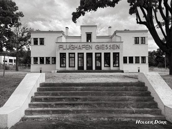 Schwarzweißaufnahme des alten Flughafengebäudes in Gießen. Das Bauwerk ist mittig ins Bild gerückt, mit klaren Linien und symmetrischem Aufbau. Über dem mittleren Eingang ist der Schriftzug „FLUGHAFEN GIESSEN“ zu lesen. Eine breite Treppe führt zum Eingang, flankiert von zwei Seitenflügeln. Die Szene wirkt ruhig und zeitlos, der Himmel ist leicht bewölkt.