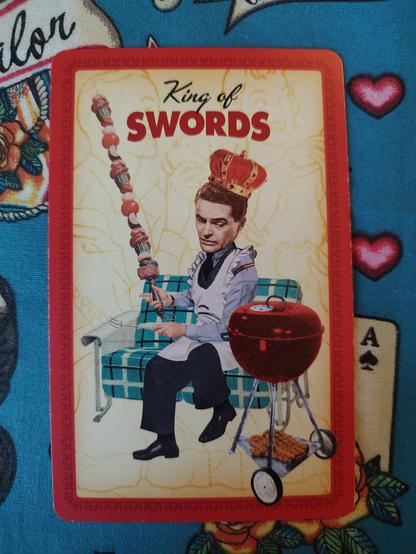 Card from the Housewives Tarot deck. 
King of Swords. 
A man, wearing a crown and a house apron, sitting on an armchair with a small barbecue grill next to him. He is holding a large kabob (the symbolic sword).
