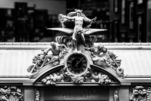 A black and white image of a close up of a miniature or model. At the Center is a decorative clock encased in an elaborate sculpture with a mostly naked man with a metal helmet that has wings (presumably the Roman god Mercury). The words "Grand Central Terminal" can be made out from the shadow below the clock sculpture.