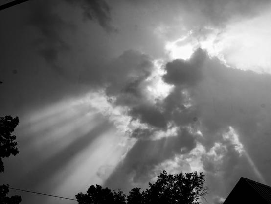 Black and white photo of dramatic clouds with sun rays streaming through gaps; silhouetted trees and the edge of a roof appear in the foreground.