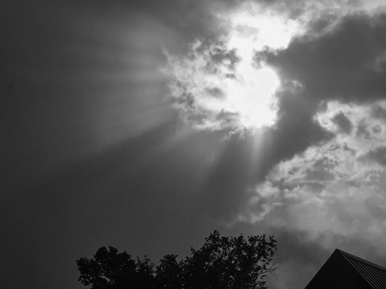 Sunlight breaks through clouds, casting dramatic rays across a dark sky. At the bottom of the black and white image, silhouettes of tree branches and a building roof are visible.