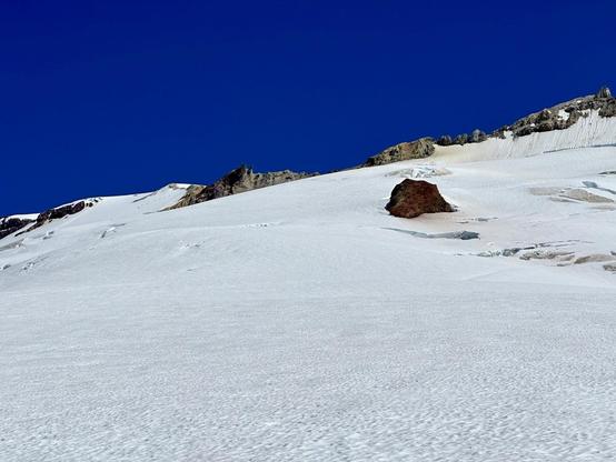 A snowy mountain landscape under a clear blue sky, featuring a prominent volcanic rock formation rising above the snow.