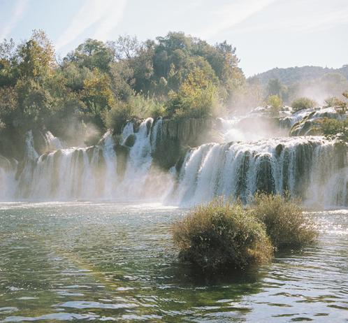 A wide waterfall flows into a river surrounded by forest. Trees fill the background, and mist floats in the air.