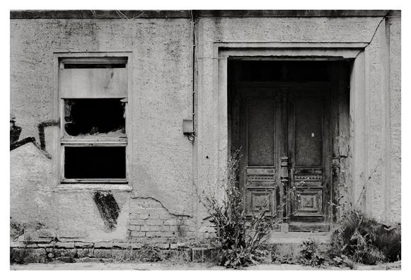Schwarz-weiß-Foto einer alten Hausfassade mit zerbrochenem Fenster links und verwitterter Doppeltür rechts. Davor wachsen Unkraut und Büsche, Putz bröckelt von der Wand.