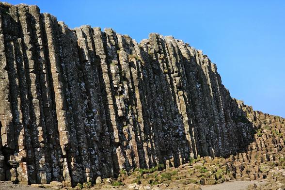 A low-angle shot of the distinctive hexagonal basalt columns of the Giant's Causeway in Northern Ireland, rising steeply against a clear, bright blue sky. The columns show varying textures and some patches of white lichen, with smaller, broken columns forming a rugged foreground at their base. The strong sunlight casts shadows that accentuate their verticality and unique formation.