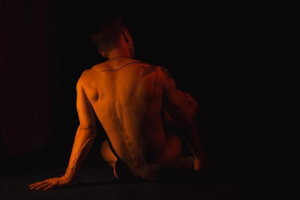 Nude individual seated on a dark floor, back facing the camera. Soft orange lighting highlights muscular back and shoulders, with a visible tattoo on the right shoulder. The person's left arm extends behind, supporting their weight.
