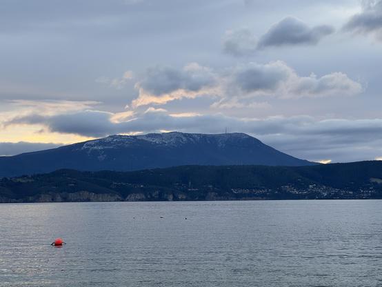 Kunanyi (Mt Wellington) seen from across the Derwent, with a blanket of grey clouds behind it, some very low behind it. The setting sun behind it has backlit some of the clouds. The mountain has snow on the higher portions. The far side of the river has the cliffs between Kingston and Taroona, with some houses from the latter visible.