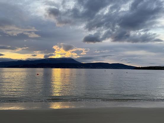 The same image of the dramatic yellows of the sun behind the grey clods over Kunanyi, but zoomed out a bit. The grey clouds over the water feel like they have motion compared to the blanket of grey cloud higher up. The water mirrors the greys or yellows from the skies above Kunanyi, but stretching all the way to the sandy shore directly in front of me