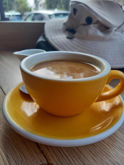 A yellow coffee cup and saucer. There is coffee with milk in the cup. A Tilley hat is behind. Cars are visible through the window.