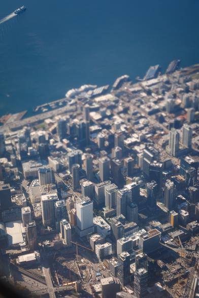 Aerial view of tall, modern buildings in central Seattle, with deep excavations at front and the waterfront then blue of Elliott Bay in the background. The centre of the photo is blurred by turbulent air from a jet engine, giving a fake tilt-shift effect of a very shallow depth of field, making the city look positively tiny.