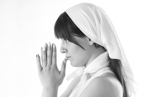 Unha muller nova de perfil con un pano branco na cabeza e as mans xuntas como para rezar