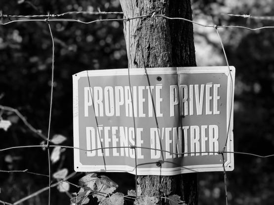 Close-up of a ‘Private property. No trespassing’ sign nailed to a fence post, behind a fence with barbed wire added, welcome... (Version françæ) Grox plan sur æn panneau 'Propriété privæ. Défense d’entrer' clouæ sur æn piquet de clôture, derrière æn grillage doublæ d’æn ligne de barbelés, bienvenue quoi…
