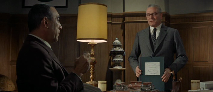 Una captura de pantalla de dos hombres en una oficina. Un hombre de piel oscura está sentado en el lado izquierdo, mirando hacia el hombre del lado derecho. Una lámpara de escritorio ilumina el centro de la imagen. El hombre de la derecha, de piel clara y gafas, está de pie detrás de un escritorio, sosteniendo una carpeta grande y oscura. La habitación está revestida de madera y detrás del hombre de la derecha hay una estantería llena de objetos.