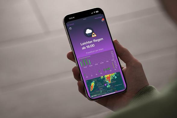 Eine Person hält ein iPhone in der Hand, auf der der Startbildschirm der Regenschirm-App mit einer Regenmeldung und einer Regenradar-Karte angezeigt wird.