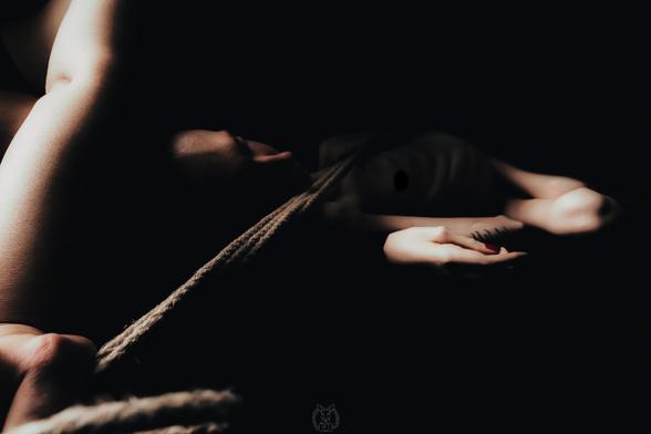 Ein Dunkel und sinnlich ausgeleuchtetes Foto von mir. Ich liege auf dem Rücken und bin nackt. Der Fokus liegt auf der Spannung des Seils, das eine Frau über meinen Körper spannt und dem Lichtspiel auf Haut, Arm und Hand. Die Szene wirkt intim, ruhig und selbstbestimmt. Eine Tätowierung und rot lackierte Fingernägel sind sichtbar – Zeichen von Individualität und Selbstinszenierung. Das Bild spielt mit Schatten und Körperlichkeit, ohne voyeuristisch zu wirken.