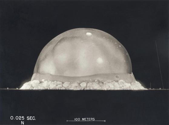 You see a black sky with a whitish, almost round uprising ball of fire on a monochrome picture of the first nuclear explosion test 80 years ago as of today. A scale in the lower part of the picture says „100m“ and the time is 0.0025sec after ignition, so diameter is ~300m at this point in time.