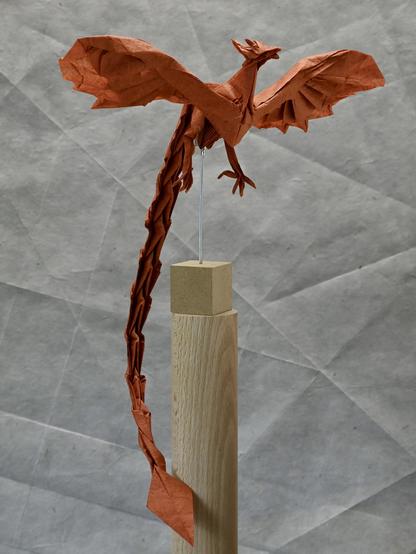 A detailed origami model of a phoenix, with spread wings and a long tail, posted on top of a wooden stand.