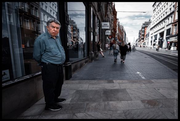 Street portrait of a man in a blue shirt standing on a pedestrianised shopping street in Birmingham, UK, with shoppers walking past and historic buildings lining both sides of the street under a cloudy sky